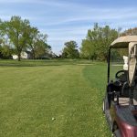 Golf cart parked on green course with expansive fairway and trees under blue sky.