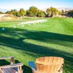 Lounge chairs on a green golf course with bright sunlight and distant hills.