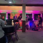 Live band performing on a stage in an indoor venue with audience seating visible.