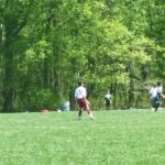Soccer game in a grassy field with trees lining the background. Players are actively engaged in play.