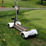 Golf push cart with putter head resting on the platform, parked on a green lawn near a golf course.