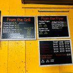 Menu board displaying food prices for grill and fryer items at a yellow food stand.