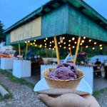 Hand holding a cone of purple ice cream in front of a green wooden pavilion at dusk.