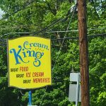 Yellow "Cream King" ice cream sign under lush green trees and utility lines.
