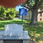 Stone monument in a grassy yard with historic buildings and trees under a blue sky.