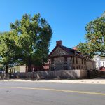 Historic stone building with red roof framed by large green trees on a sunny day.