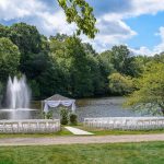 Outdoor wedding ceremony setup by a pond with trees and fountain in the background.