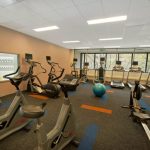 Gym interior with various cardio machines and large windows overlooking trees.