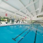 Indoor swimming pool with bright white ceiling and large windows overlooking a green outdoor area.