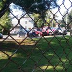 Chain-link fence view of parked cars and a house on a sunny day.