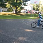 Cyclist riding a black bike on an asphalt court with trees and buildings in the background.