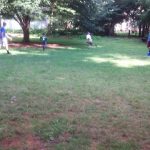 People playing sports on a grassy field surrounded by trees under bright daylight.