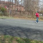 Young person plays basketball on an outdoor court with trees and buildings in the background.