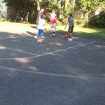 Children playing soccer on an outdoor asphalt court under trees.