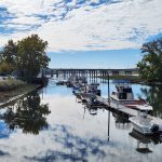Boats moored under a bridge on a calm waterway with blue sky and clouds.