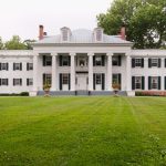 White neoclassical mansion with columns set against a lush green lawn and trees.