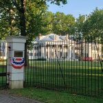 Historic white mansion behind a black wrought iron fence, surrounded by lush green trees.