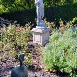 Stone statue of a figure in a garden setting with lush greenery and shrubs.