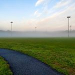 Foggy sports field with a dark pathway leading across the green grass at sunrise.