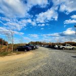 Gravel parking lot under a bright blue, cloudy sky with trees in the background.