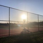 Backlit tennis court through chain-link fence at sunset or sunrise.