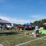 Outdoor gathering with tents, cars, and people on a grassy field under a blue sky.