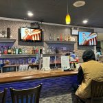 Bar interior with patrons seated at the counter, featuring TVs and a tiled bar front.
