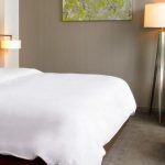 Bright hotel room with a large white bed, modern lamp, and neutral decor.