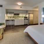 Modern hotel room with living area, kitchenette, and large white bed.