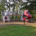 Children play on a colorful playground structure surrounded by trees in a park setting.