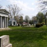 Historic cemetery scene with classical mausoleum, gravestones, and trees under a bright sky.