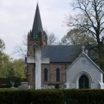 Gothic church with spire and white cross, framed by trees in a cemetery setting.