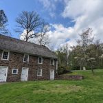 Stone farmhouse on a grassy hill under a partly cloudy blue sky with trees surrounding it.