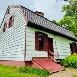 White clapboard house with dark roof and red trim, set against green trees.