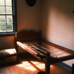Wooden bed in a sparsely furnished room with sunlight streaming through a window.