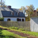 White farmhouse with dark roof behind a wooden picket fence and small shed.