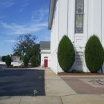 White classical building with tall window and green shrubs on a sunny day.