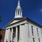 White neoclassical church with a tall steeple under a bright blue sky.