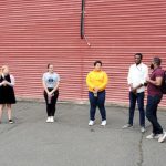 Group of people gathered outdoors in front of a large red corrugated metal building.