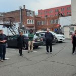 Group of people gathered outdoors in a parking lot near brick and modern buildings.