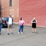 Group of people standing outside a red industrial building, appearing to be in a meeting or gathering.