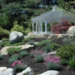 White gazebo nestled in a lush, rocky garden setting with surrounding greenery.