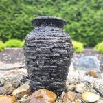 Dark, stacked stone garden urn sits on gravel with green hedge background.