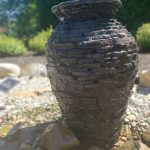 Rustic, dark stone pot sits on a rocky beach with green foliage in the background.