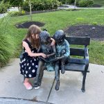 Bronze statue of three figures seated on a park bench outdoors.