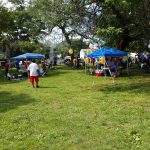Outdoor community gathering with tents, people socializing, and a barbecue setup on green lawn.