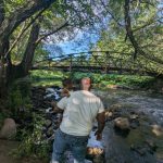 Father and child walk across a rocky stream under an arched bridge on a sunny day.