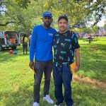 Two men stand outdoors in a grassy park under leafy trees.