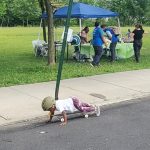 Child wearing helmet skateboarding on asphalt near a park event tent.