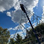 Child climbs a large, intricate spiderweb-like climbing structure under a blue sky with white clouds.